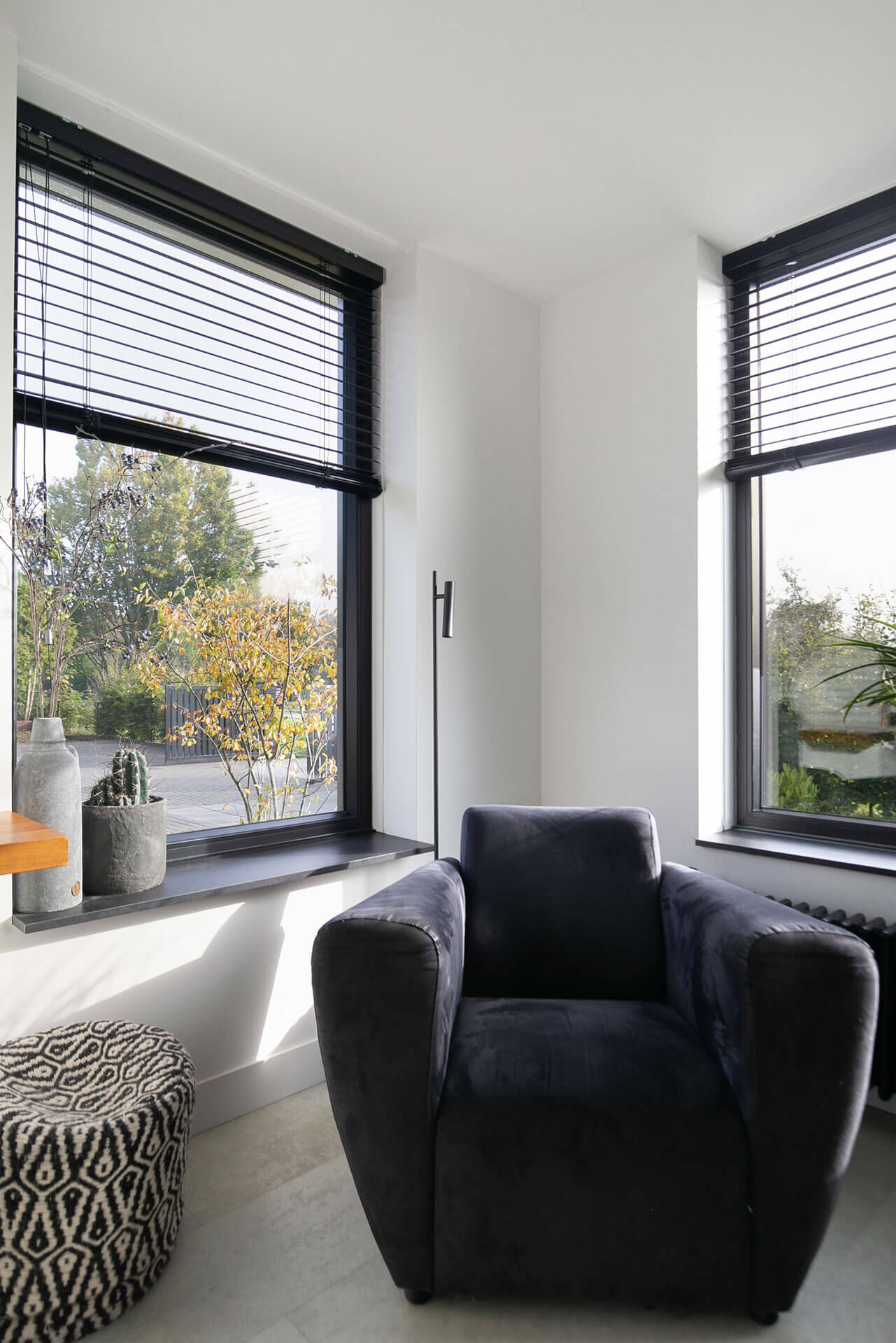Zwarte kozijnen met houten jaloezieën in een lichte woonkamer met fauteuil en uitzicht op de tuin