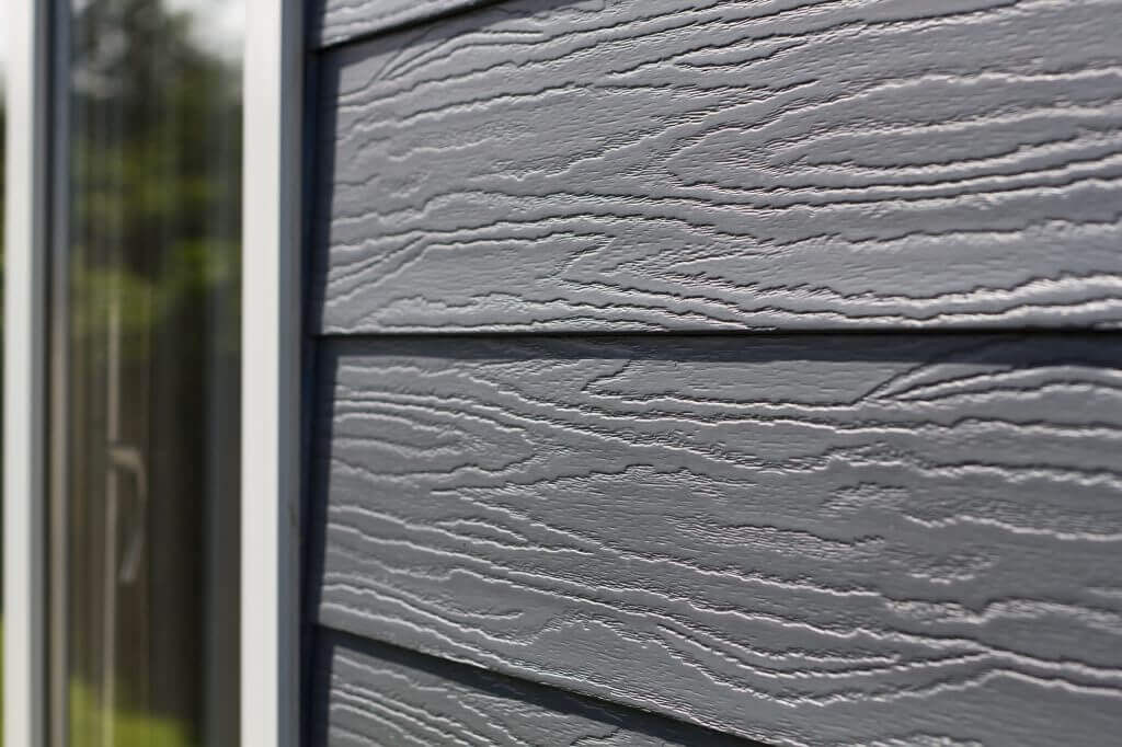 Close-up van grijze gevelbekleding met houtnerfstructuur naast een wit kozijn
